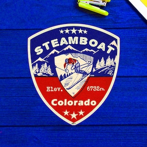 May include: A vintage-style red, white and blue shield-shaped sticker with the text "STEAMBOAT" in white letters. The sticker features a skier in blue and white clothing, mountains, and trees. The text "Elev. 6732 ft. Colorado" is printed below the skier.