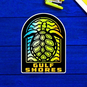 Puede incluir: Una pegatina con un diseño de tortuga marina y el texto "GULF SHORES". La tortuga es negra con detalles amarillos y verdes, sobre un fondo azul y amarillo. La pegatina tiene un borde negro.