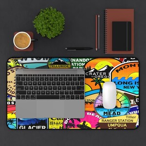 May include: A colorful desk setup featuring a laptop, mouse, and a vibrant mousepad adorned with travel-themed stickers. A cup of coffee, a small plant, and a notebook are also present. The mousepad has stickers from Crater Lake, Long Island, and other locations.