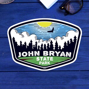 May include: A white sticker with a blue, green, and white design. The sticker features a stylized image of a forest with a bird flying over it. The text "John Bryan State Park" is printed in white on the sticker.
