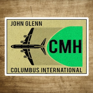 May include: A vintage-style sticker with a black airplane flying over a green circle with the text "CMH" in black. The text "JOHN GLENN COLUMBUS INTERNATIONAL" is printed in black at the top and bottom of the sticker.