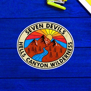 May include: A vintage-style sticker with a mountain scene and the text "Seven Devils Hells Canyon Wilderness". The sticker has a red, orange, yellow, and blue color scheme.