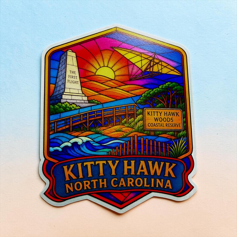 May include: A colorful sticker featuring a stained-glass style illustration of Kitty Hawk, North Carolina. It depicts the Wright Brothers National Memorial with the text The First Flight, a hang glider, a wooden boardwalk, and a Kitty Hawk Woods Coastal Reserve sign.