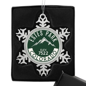 May include: Silver snowflake ornament with a green circle in the center. The circle says "Estes Park, Colorado" and "Elev. 7522" in white text.
