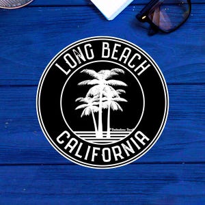 May include: A black and white circular sticker featuring the text "LONG BEACH CALIFORNIA" around a palm tree design. The sticker is placed on a blue wooden surface, with office supplies visible in the background.