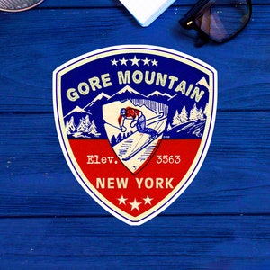 Peut inclure: Un autocollant de style vintage avec un design de bouclier bleu et rouge. L'autocollant représente un skieur sur une montagne avec le texte "Gore Mountain" et "Elev. 3563 NEW YORK" en lettres blanches.