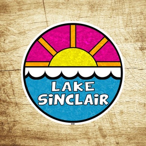 Könnte beinhalten: Ein runder Aufkleber mit einem blauen, weißen und gelben Sonnenuntergang über einem Gewässer. Der Text "LAKE SINCLAIR" ist in weißen Buchstaben auf blauem Hintergrund geschrieben.