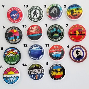 May include: Fifteen round, colorful buttons with various designs and text. The buttons feature images of palm trees, mountains, a surfer, a rocket, a Bigfoot, a lake, a sunset, and more. The text on the buttons includes "Space Camp", "Gone Squatchin", "Maui Surf Riders", "Malibu Beach", "Venice Beach", "Lake Tahoe", "Huntington Beach", "Santa Cruz Surf Club", "Great Smoky Mountains National Park", "The Mountains Are Calling", "Explore", "Yosemite", "Miami Beach", "Beach Life", and "Hike More Worry Less".