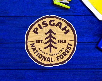 May include: A brown and tan woodgrain sticker with the text "Pisgah National Forest" in a circle around a stylized pine tree. The text "Est. 1916" is inside the circle. The text "North Carolina" is below the tree.