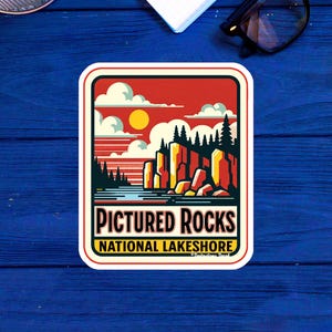 May include: A vintage-style sticker with a red, orange, and yellow sunset scene over a lake with green trees and red rock cliffs. The text reads "Pictured Rocks National Lakeshore".