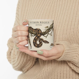 May include: A white ceramic mug featuring a detailed illustration of a Ball Python, with the text "PYTHON REGIUS" and "Ball Python". The snake is depicted in brown and tan tones, coiled around green vines.