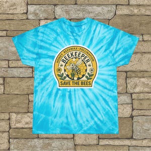 Può includere: Una maglietta turchese tie-dye con una grafica circolare raffigurante un'ape, un favo e dei fiori. La grafica include le parole "BEEKEEPER" e "SAVE THE BEES", insieme a testo su miele, cera d'api e impollinazione.