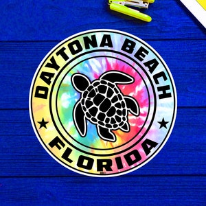 May include: A round sticker with a tie-dye background and a black silhouette of a sea turtle. The sticker says "Daytona Beach Florida" in a white circle around the turtle.