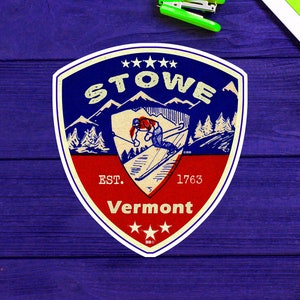 Op de afbeelding: Een sticker in vintage stijl met een blauw, rood en wit kleurenschema. Het toont een skiër in een rood en wit pak, een bergketen en de tekst "STOWE EST. 1763 Vermont".