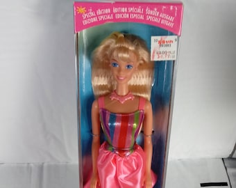 May include: A Barbie doll in a clear plastic box. The doll has blonde hair and is wearing a pink dress with a rainbow-striped top. The box reads "Children's Day KinderTags Barbie".