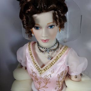 May include: A porcelain doll with brown hair styled in curls and a gold tiara. The doll wears a pink dress with gold trim and a jeweled necklace. The doll has blue eyes and is packaged in a box.