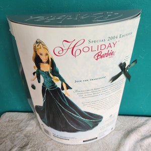 May include: Special 2004 Edition Holiday Barbie doll in a dark teal velvet gown.  The box features the doll in her elegant gown, along with text highlighting the special edition and the tradition of Holiday Barbie dolls. The back of the box includes details about the doll and the tradition.  The doll is shown holding a small purse. The box is silver and white.
