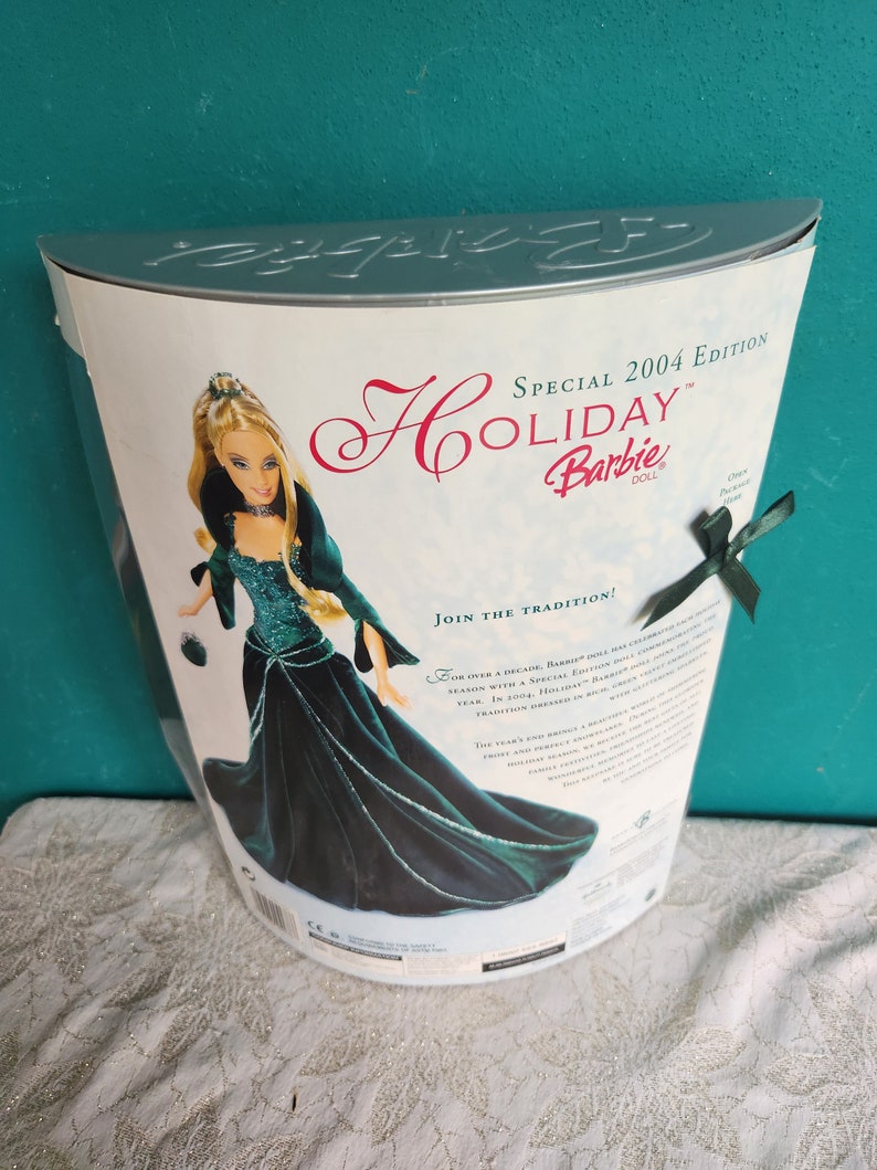 May include: Special 2004 Edition Holiday Barbie doll in a dark teal velvet gown.  The box features the doll in her elegant gown, along with text highlighting the special edition and the tradition of Holiday Barbie dolls. The back of the box includes details about the doll and the tradition.  The doll is shown holding a small purse. The box is silver and white.