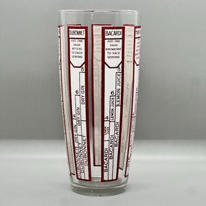 Peut inclure: Un shaker &agrave; cocktail en verre transparent avec des &eacute;tiquettes rouges et blanches pour la pr&eacute;paration de cocktails. Les &eacute;tiquettes comprennent les marques Dubonnet et Bacardi, ainsi que des instructions pour ajouter des ingr&eacute;dients tels que du gin sec, du jus de citron et de la grenadine.