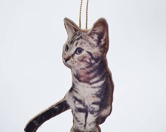 May include: A cat-shaped ornament with a gold string for hanging. The ornament features a detailed image of a tabby kitten with gray and brown stripes. The cat is sitting with its tail extended. The background is a soft, neutral color.
