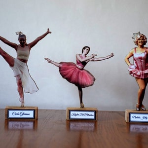 Peut inclure: Trois figurines en bois de danseuses, chacune avec une pose et une tenue différentes. Les figurines sont sur des socles en bois avec des noms imprimés dessus : "Carla Gleason", "Kayla DeMattia" et "Natalie Jelly Victoria".