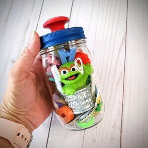 May include: A clear glass jar with a blue lid and red handle, filled with colorful toys and a green Oscar the Grouch figurine. The jar is held by a person with a pink watch.