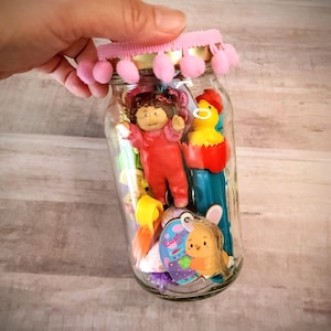 May include: A clear glass jar filled with small toys and trinkets, including a red doll, a yellow chick, and an Easter Day tag. The jar is topped with a pink pom-pom trim and held by a hand.