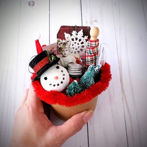 May include: A miniature Christmas-themed diorama featuring a snowman with a black top hat, a vintage photo, miniature trees, and a snowflake tag. The diorama is nestled in a red fuzzy container, creating a festive holiday scene.