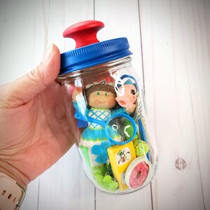 May include: A clear glass jar with a blue lid and red knob, filled with small toys and figurines. The jar contains a doll, a pink-faced figure, a magnifying glass, a yellow block with a cartoon character, and a pink donut.