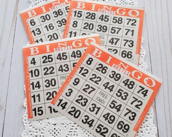 May include: Four vintage bingo cards with orange borders and black numbers are arranged on a white paper doily. The cards display the word "BINGO" at the top and have various numbers in a grid format. The cards are printed in the USA.