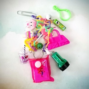 May include: A collection of colorful plastic charms and keychains, including a pink purse, a pink ice cream cone, a pink and purple drink, a green bell, a green bottle, and a rainbow chain.