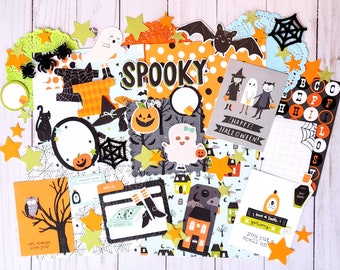 May include: A collection of Halloween-themed paper cutouts and embellishments. Includes the word "SPOOKY", ghosts, bats, pumpkins, stars, and the text "Happy Halloween!". The color palette features orange, black, white, and green.