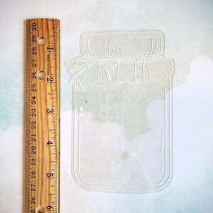 May include: A white outline of a mason jar with a lid and a rope tied around the lid. The jar is about 3 inches tall.
