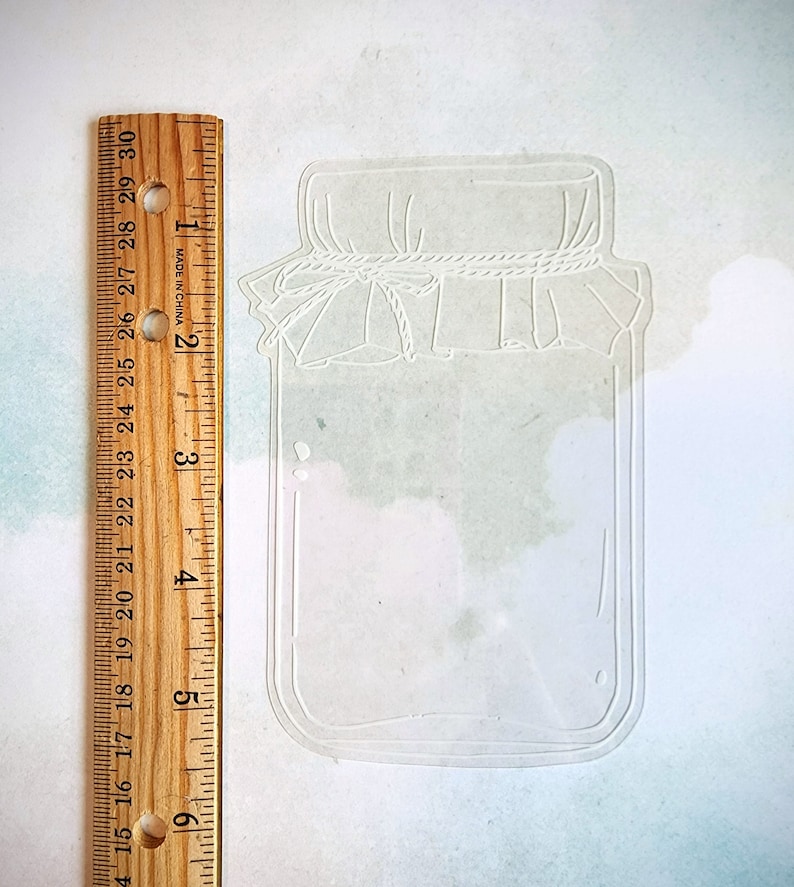 May include: A white outline of a mason jar with a lid and a rope tied around the lid. The jar is about 3 inches tall.