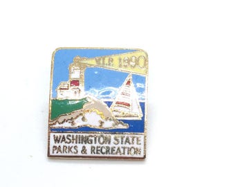 May include: A rectangular pin featuring a lighthouse, sailboat, and the text "V.I.P. 1990." The pin also includes the words "WASHINGTON STATE PARKS & RECREATION" in a gold-colored frame. The background is blue with green and white accents.