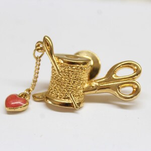 May include: A gold-coloured brooch depicting a thread spool with a needle, scissors, and a dangling red heart charm. The brooch is likely made of metal and has a detailed, textured design.