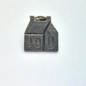 May include: A silver-toned metal pin shaped like a house with three windows. The pin has a simple design and is a great addition to any collection.