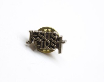 May include: A silver pin with the words "Jesus First" in a stylized font. The pin has a gold backing and a cross.