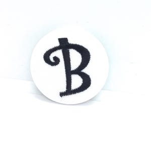 May include: A white, circular patch with a black, stylized letter "B" embroidered in the center. The letter has a decorative, swirling top and a simple, straight bottom.