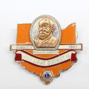 May include: A silver and orange enamel pin featuring a portrait of Benjamin Harrison, the 23rd President of the United States. The pin also includes the text "Honolulu", "Benjamin Harrison", "Ohio Lions District 13", and "1983".
