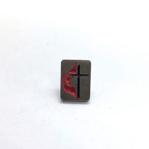 May include: A rectangular metal lapel pin with a brushed silver finish. The pin features a black cross centered on the right side and a stylized red flame design extending from the left side of the cross, creating a distinct contrast against the metal.
