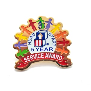 May include: A gold-coloured pin with a circular design. The pin features the words "HEAD START 5 YEAR SERVICE AWARD" in red and white. The design includes colourful stick figures and a wheelchair symbol.