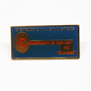 May include: Gold-colored pin with a blue background. The pin features a key with the word "Commitment" and the text "District 8 Toastmasters" at the top. The text "Commitment is the key to success" is written on the pin.