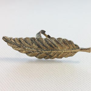 May include: A metallic ring featuring a long, horizontal leaf-shaped design. The surface of the leaf has a textured, weathered bronze finish with visible veins and a pointed tip. The band of the ring is visible behind the center of the leaf.