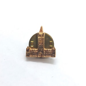 May include: A gold-coloured metal lapel pin featuring a detailed architectural relief of a building with a central tower and a pointed spire. The design includes a circular backing plate with a textured surface and a pin mechanism on the reverse side.