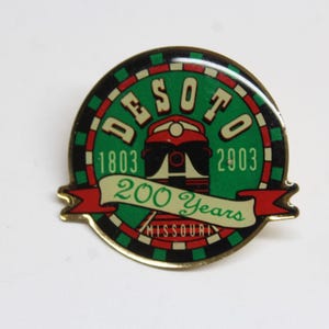 May include: A green and red circular pin with a gold border. The pin features a black and white illustration of a train engine with the text "DESOTO 1803 2003" and "200 Years MISSOURI" around the engine.