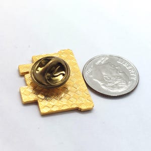 May include: A gold-coloured lapel pin with a geometric design and a textured surface. The pin has a butterfly clutch backing. A US dime is next to the pin for size comparison.