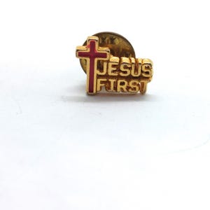 May include: A gold-toned metal lapel pin featuring a red cross and the raised text JESUS FIRST. The pin has a metallic finish and a standard clutch backing, displaying a clear religious message in a compact, three-dimensional design.