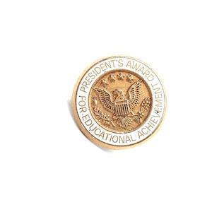 May include: A gold-coloured President's Award for Educational Achievement pin. The pin features an eagle emblem at the centre, surrounded by text. The text reads "PRESIDENT'S AWARD FOR EDUCATIONAL ACHIEVEMENT".