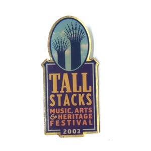 May include: A rectangular pin with a gold border and the words "TALL STACKS" in gold lettering on a purple background. The pin also features the text "MUSIC, ARTS & HERITAGE FESTIVAL 2003" and an image of two tall structures.